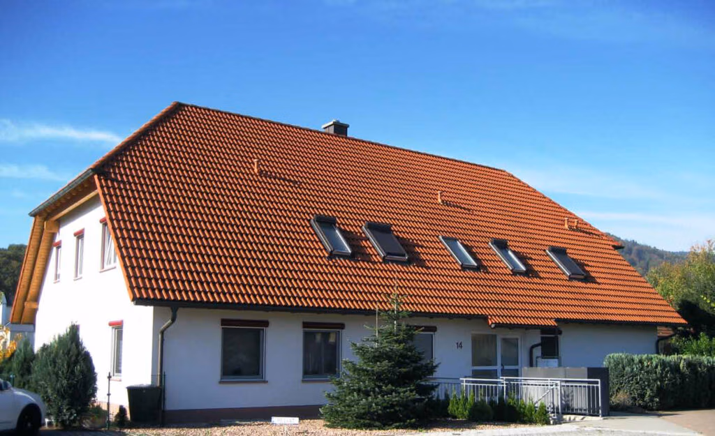 Ein Wohnhaus mit weißer Fassade, rotem Ziegeldach und mehreren Dachfenstern, umgeben von Bäumen, einem kleinen Vorgarten und einem blauen Himmel im Hintergrund.